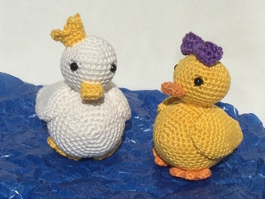 Zwei gehäkelte Enten-Amigurumi, links weiß mit gelber Krone, rechts gelb mit lila Schleife auf blauem Untergrund