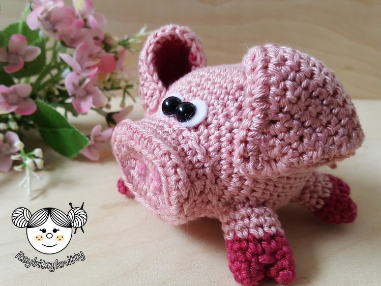 Rosa gehäkeltes Schwein mit schwarzen Augen und dunklen Hufen auf hellem Holz, mit rosa Blüten im Hintergrund.