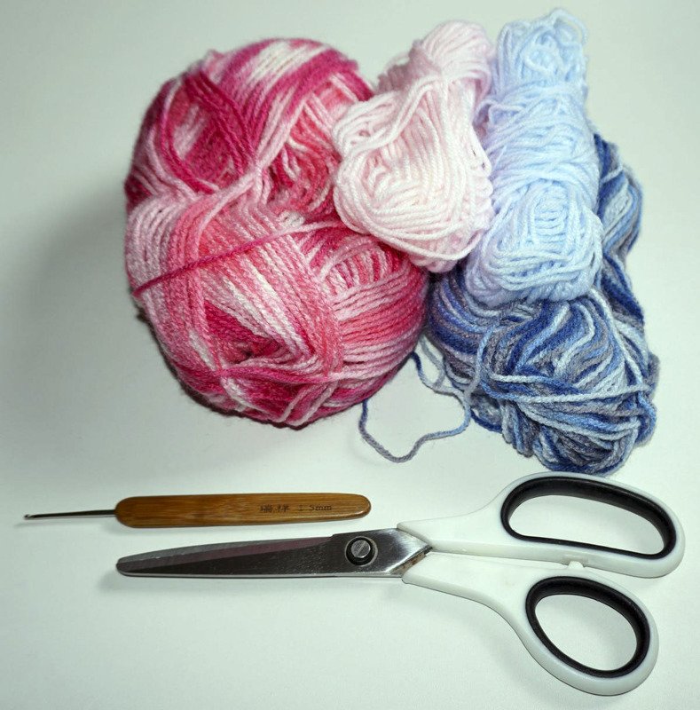 Assorted pink and blue yarn skeins with a wooden-handled crochet hook and white-handled scissors on a white background