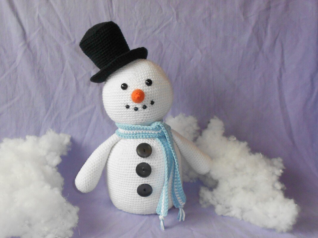 Gehäkelter weißer Schneemann mit schwarzem Zylinderhut, orangefarbener Nase, blau-weiß gestreiftem Schal und drei schwarzen Knöpfen vor lila Hintergrund