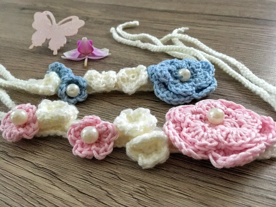 Gehäkeltes Stirnband mit blauen, rosa und cremefarbenen Blumen und Perlen auf Holzuntergrund, daneben eine Papier-Schmetterlingsfigur und eine Orchideenblüte.