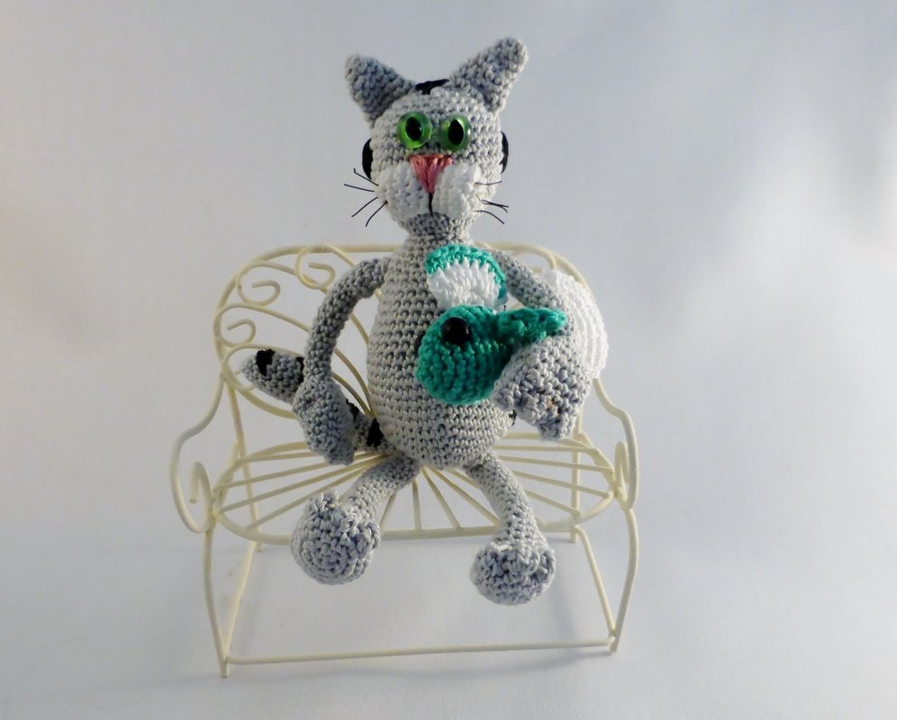 Gehäkelte graue Amigurumi-Katze mit grünen Augen und rosa Nase auf einer kleinen weißen Metallbank, die einen türkisfarbenen gehäkelten Fisch hält.