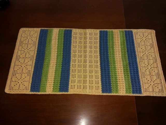 Gehäkelter rechteckiger Tischläufer in Beige mit blauen und grünen Streifen und Lochmuster auf dunklem Holztisch.