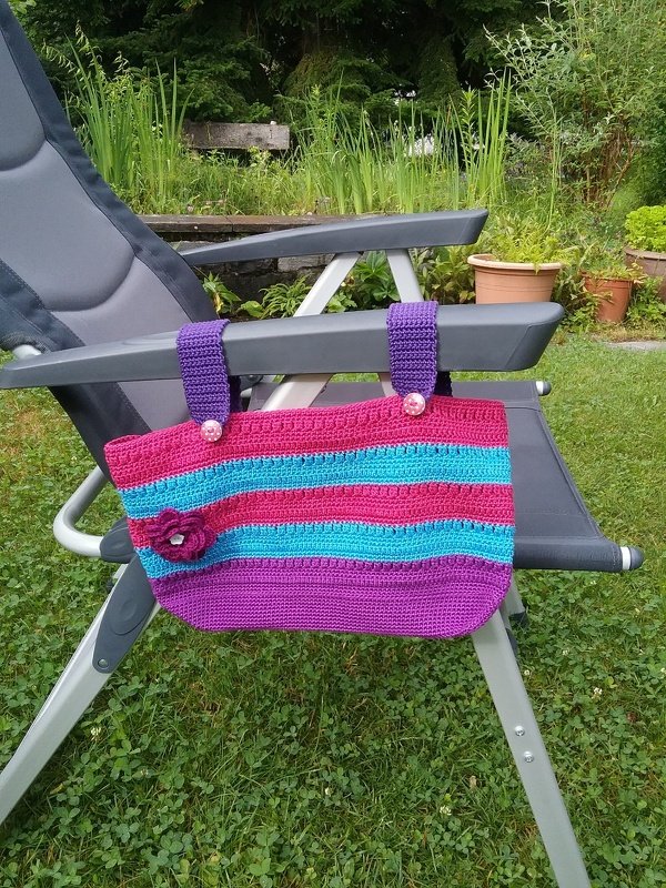 Gehäkeltes Utensilo in Pink, Türkis und Lila mit Schlaufen und Knöpfen, an einer Gartenliege befestigt