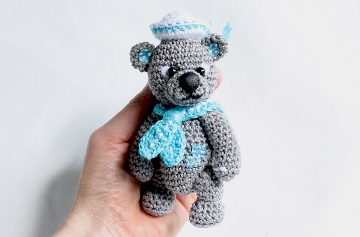 Hand hält kleinen grauen gehäkelten Teddybär mit weißem Matrosenhut und hellblauem Schal vor hellem Hintergrund.