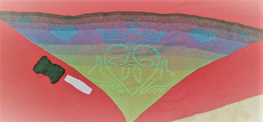 Gehäkeltes Dreieckstuch in Blau-, Violett- und Grüntönen mit Kronen- und Herzmotiv, Garnknäuel und Etikett daneben