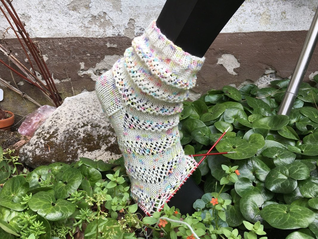 Unfertige gestrickte Socke mit Ajourmuster in hellem Garn und Farbpunkten, an einem Bein getragen. Sichtbar sind Stricknadeln und eine grüne Pflanzenumgebung.