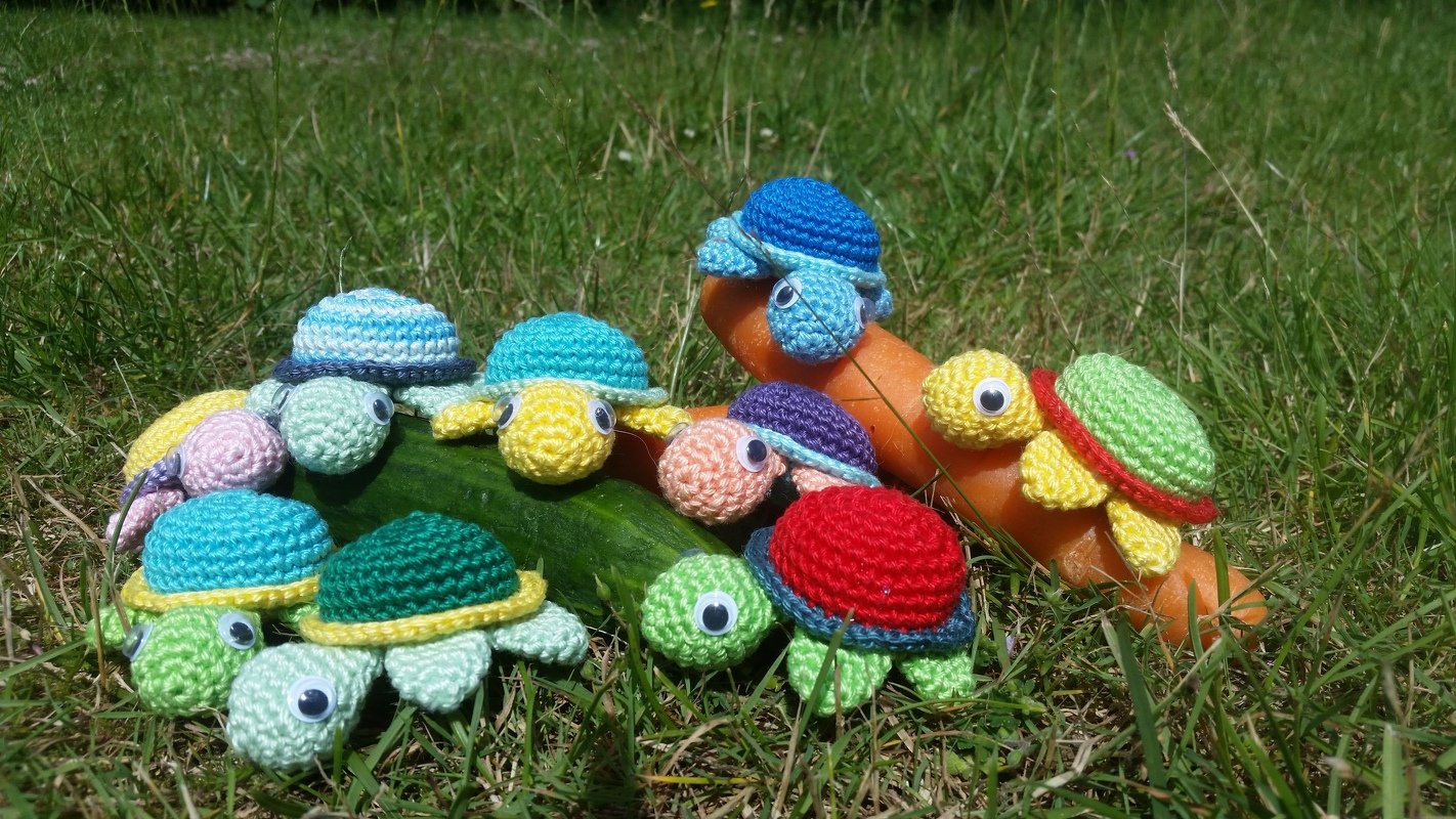 Mehrere kleine gehäkelte Amigurumi-Schildkröten in verschiedenen Farben auf Gras neben Gurke und Karotten