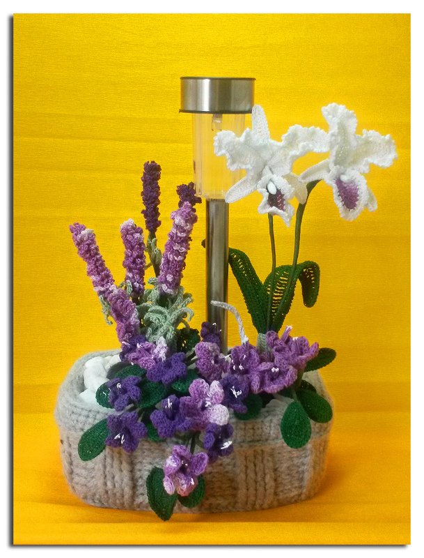 Gehäkelte Lavendel-, Veilchen- und Orchideenblüten in grauem Häkelübertopf mit Solarlampe vor gelbem Hintergrund