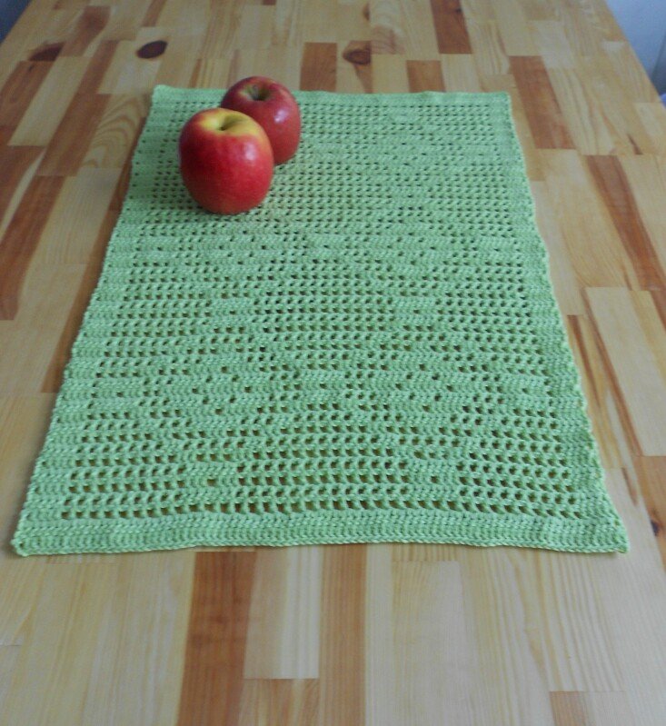 Hellgrüner gehäkelter Tischläufer mit Filetmuster auf einem Holztisch, darauf zwei rote Äpfel.