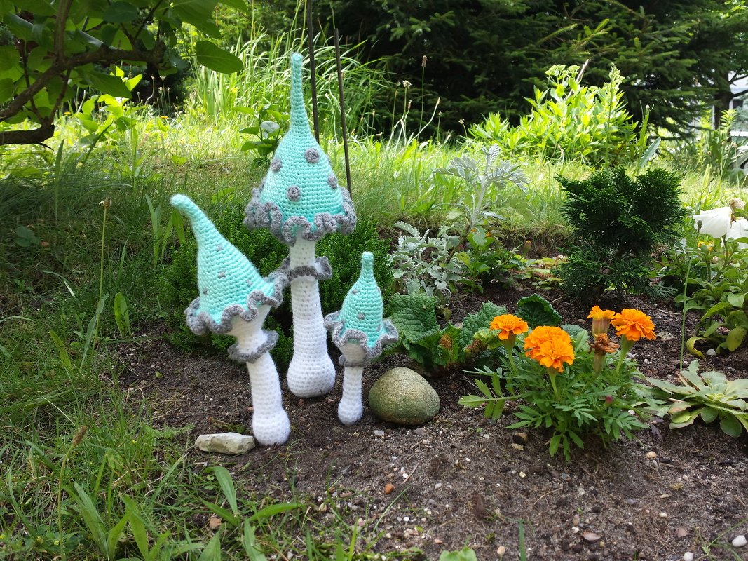 Drei gehäkelte Zierpilze in Mintgrün, Grau und Weiß zwischen Pflanzen im Garten.