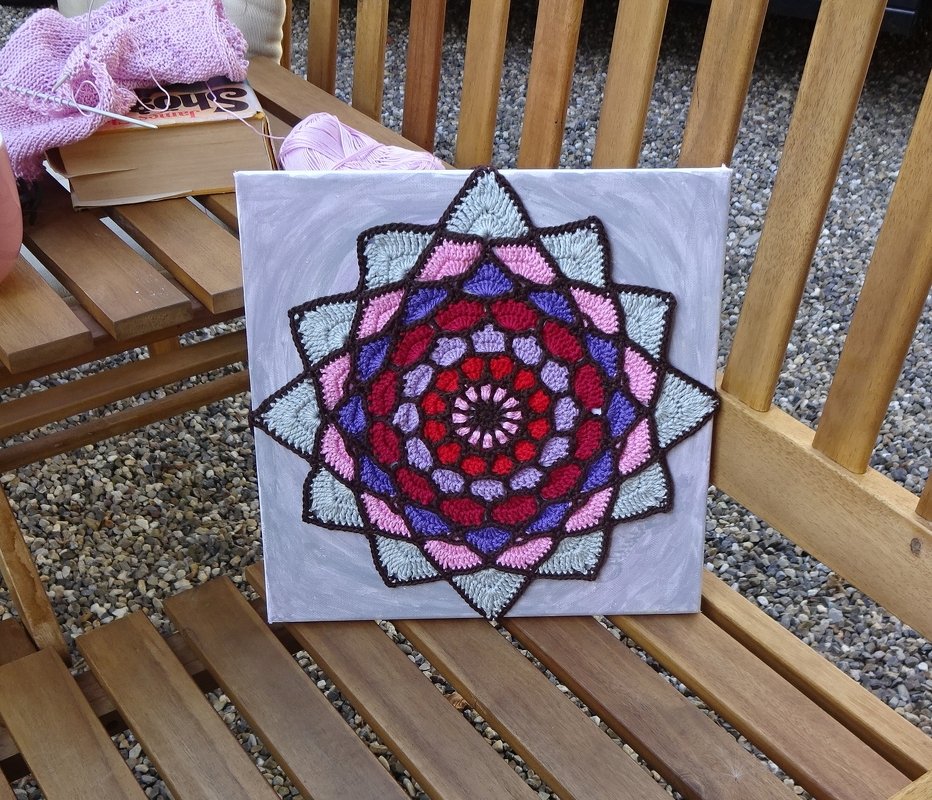 Gehäkeltes Mandala in Rot, Rosa, Violett und Hellblau auf quadratischer Leinwand auf einer Holzbank mit Wollknäuel