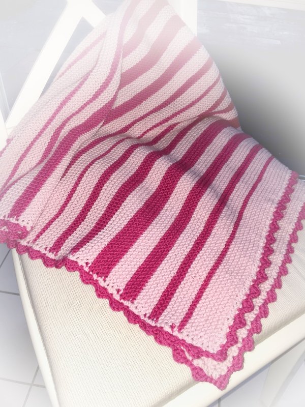 Gestrickte Babydecke in Hell- und Dunkelrosa mit diagonalen Streifen und gewellter Kante auf einem hellen Stuhl.