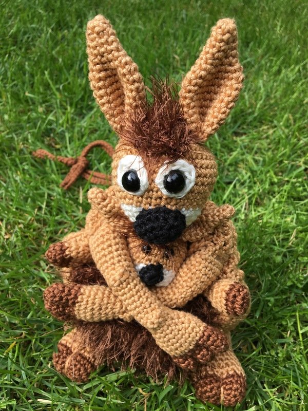 Zwei braune, gehäkelte Amigurumi-Tierfiguren, eine größere umarmt eine kleinere, sitzen auf grünem Gras.