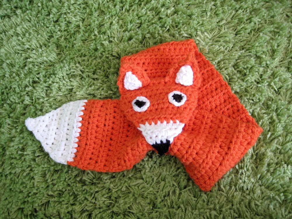 Crocheted orange fox scarf with white ears, snout, and tail tip, featuring black eyes and nose on a green textured surface.