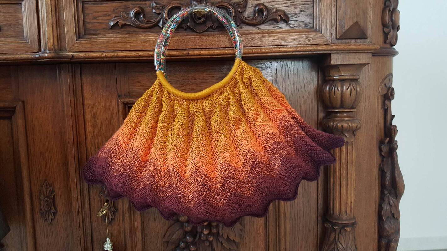 Gehäkelte Tasche in Gelb, Orange und Burgunderrot mit transparentem Ringhenkel, gefüllt mit bunten Perlen, hängend an einem geschnitzten Holzschrank