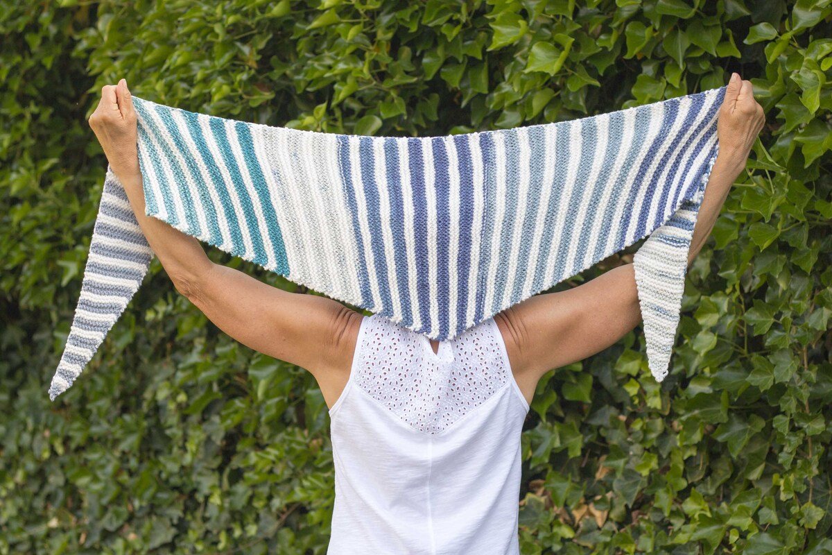 Person hält blau-weiß gestreiftes gestricktes Dreieckstuch vor grüner Hecke