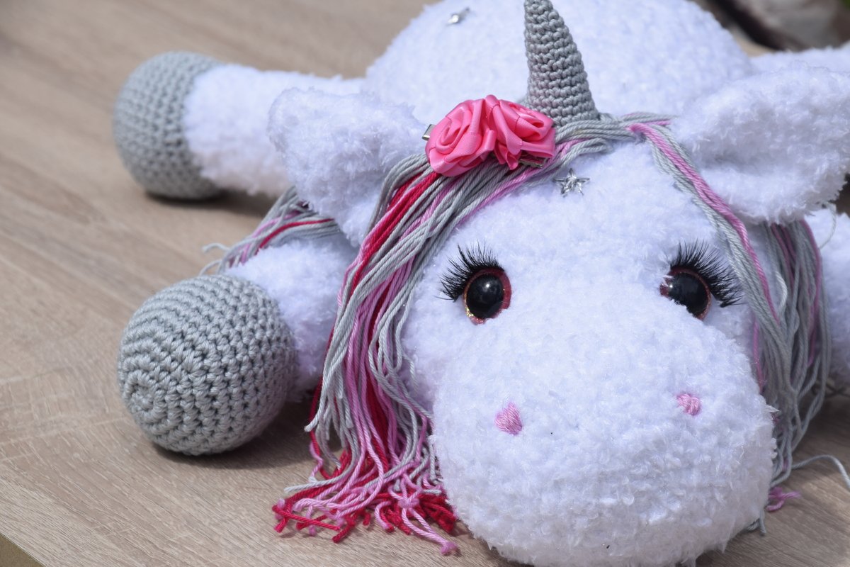 Liegendes gehäkeltes Einhorn-Amigurumi mit weißem, flauschigem Körper, grauem Horn und Hufen sowie pink-grauer Mähne auf Holzoberfläche.