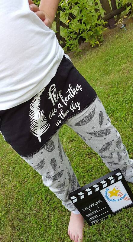 Hosenbund mit schwarzem Einsatz zeigt eine weiße Feder und den Schriftzug "fly like a feather in the sky". Die hellgrauen Hosenbeine sind mit einem wiederholten Federmuster bedruckt. Eine Person steht barfuß auf Gras; eine Filmklappe liegt am Boden.