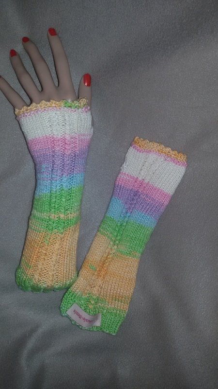 Zwei gestrickte fingerlose Armstulpen mit pastelligen Streifen, eine an einer Schaufensterhand, eine liegend.