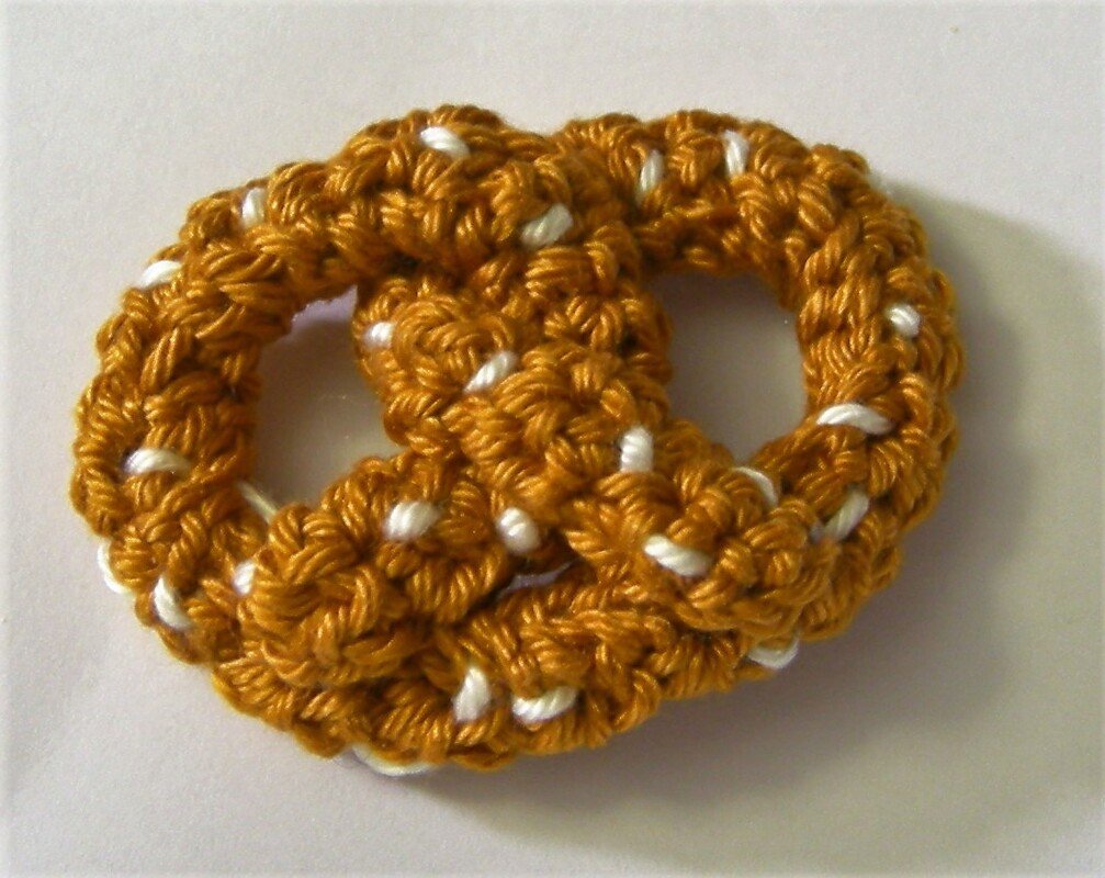 Gehäkelte goldbraune Brezel mit weißen Stichen auf hellem Untergrund.