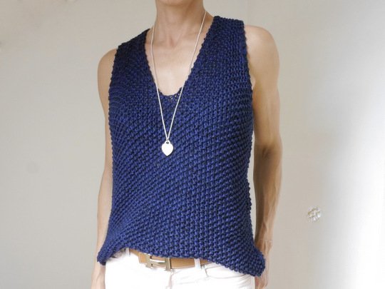 Person trägt ein dunkelblaues, gestricktes ärmelloses Top mit V‑Ausschnitt und markantem Strukturmuster, kombiniert mit weißer Hose und einer Herz‑Halskette.