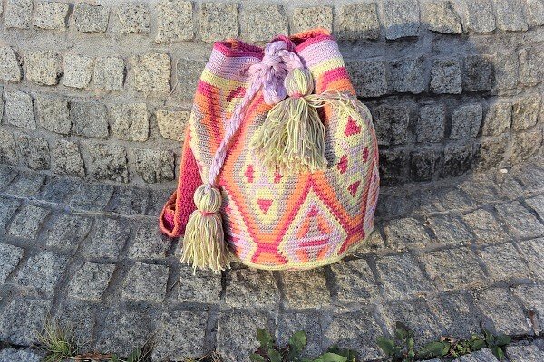 Gehäkelte Umhängetasche in Pink, Orange und Gelb mit geometrischem Muster und Quasten auf einer Steinmauer.