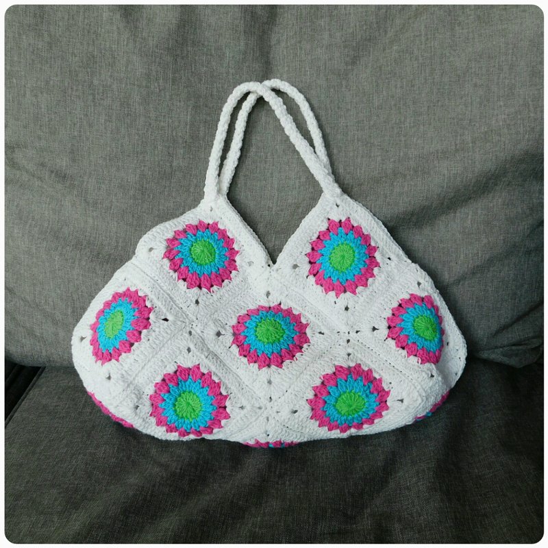 Weiße Henkeltasche aus Granny Squares mit grünen, türkisen und pinken Blüten auf grauem Hintergrund.