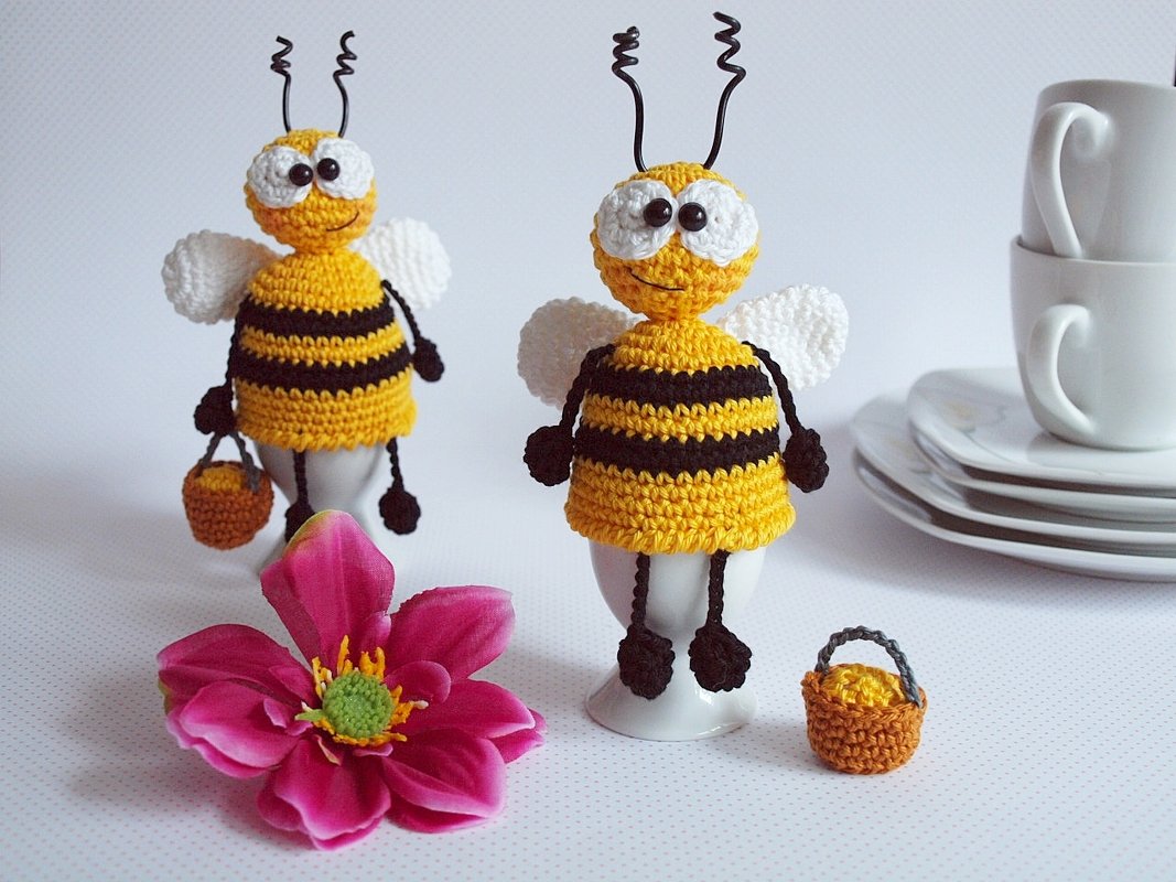 Zwei gehäkelte Bienen-Eierwärmer in Gelb und Schwarz auf Eierbechern, davor eine pinke Blüte und ein kleines gehäkeltes Körbchen, im Hintergrund gestapelte Tassen und Teller