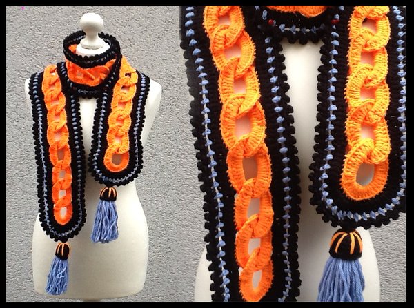 Langer gehäkelter Schal mit orangefarbenen Kettengliedern, schwarzer Umrandung und hellblauen Quasten auf einer Schneiderpuppe