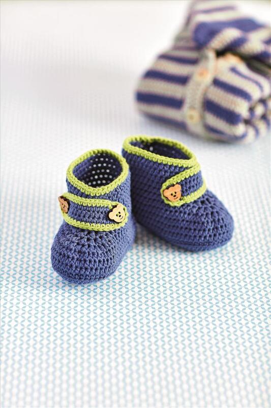 Gehäkelte dunkelblaue Babyschuhe mit hellgrünem Rand und kleinen Bärenknöpfen, auf gemustertem Untergrund mit gestreifter Decke im Hintergrund