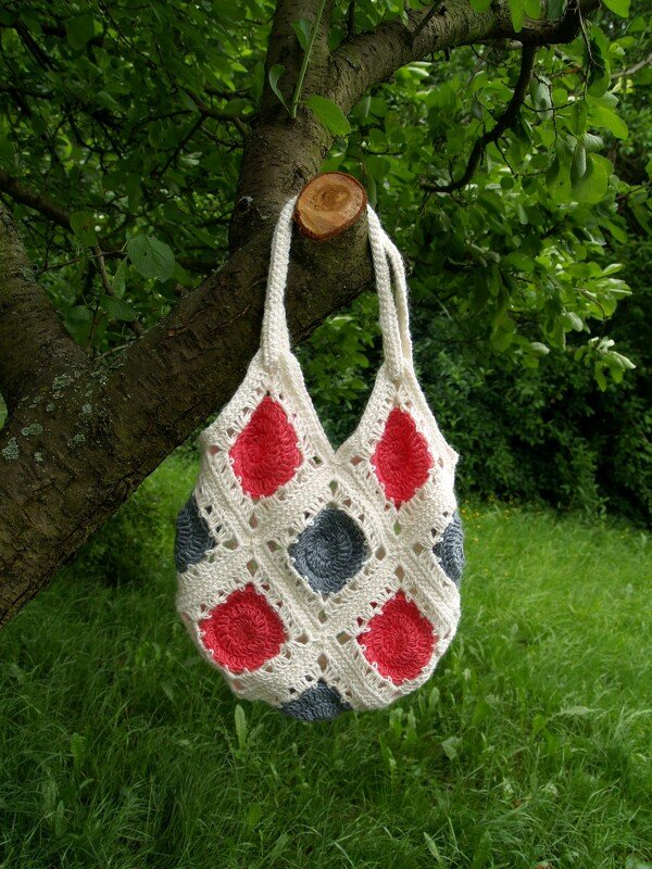 Gehäkelte Henkeltasche aus Granny Squares in Creme mit roten und grauen Mittelpunkten hängt an einem Baum