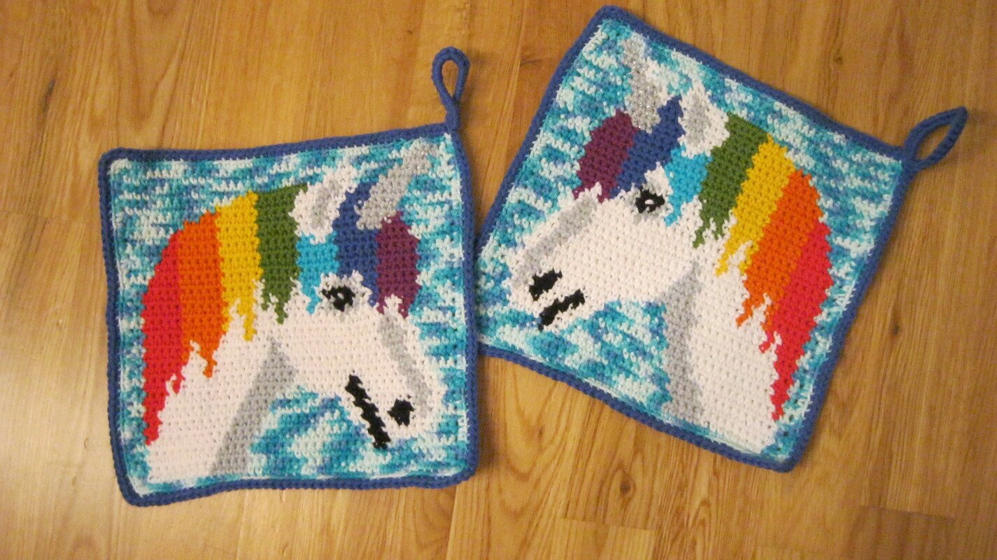 Zwei quadratische gehäkelte Topflappen mit Einhornkopf und Regenbogenmähne auf türkisgemustertem Hintergrund