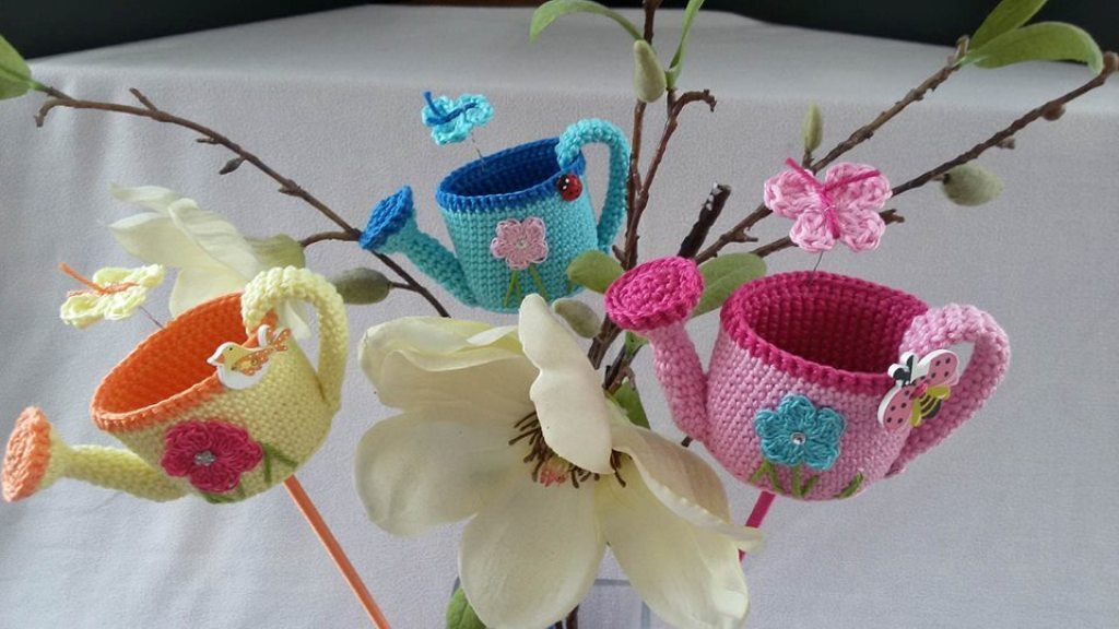 Drei bunte gehäkelte Gießkannen in Gelb, Türkis und Rosa, verziert mit kleinen Applikationen, arrangiert mit Zweigen und weißen Blüten.