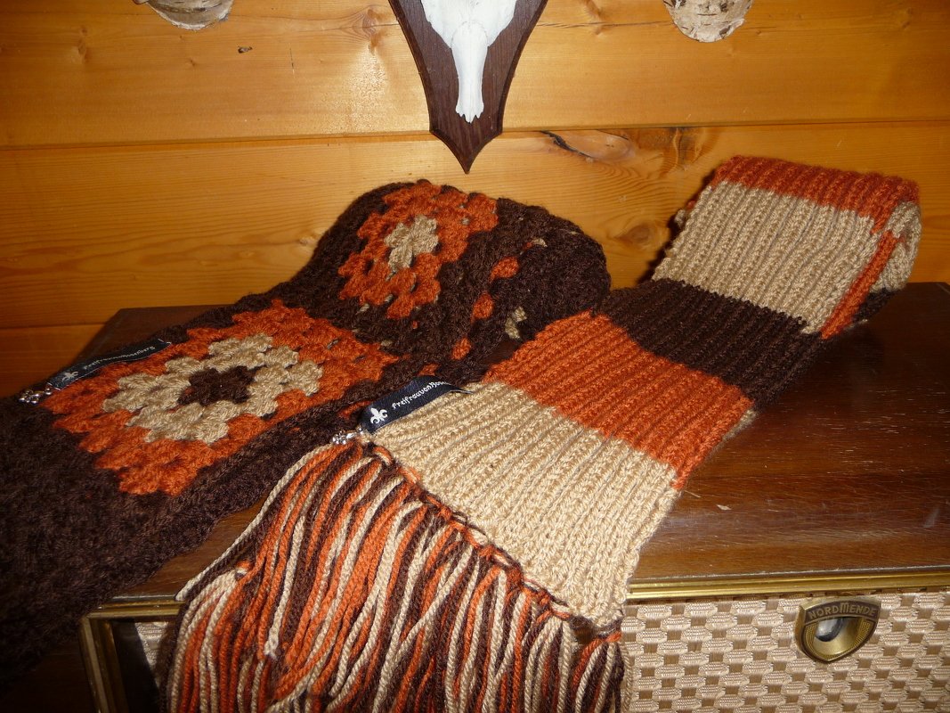 Zwei Schals auf Holz: ein gehäkelter Granny‑Square‑Schal in Braun und Orange sowie ein gestrickter Streifenschal mit Fransen in Beige, Braun und Orange.