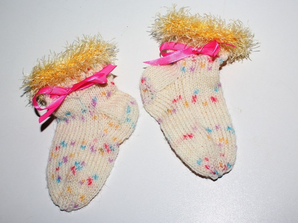 Zwei cremefarbene handgestrickte Babysocken mit bunten Sprenkeln, gelben flauschigen Bündchen und rosa Satinschleifen