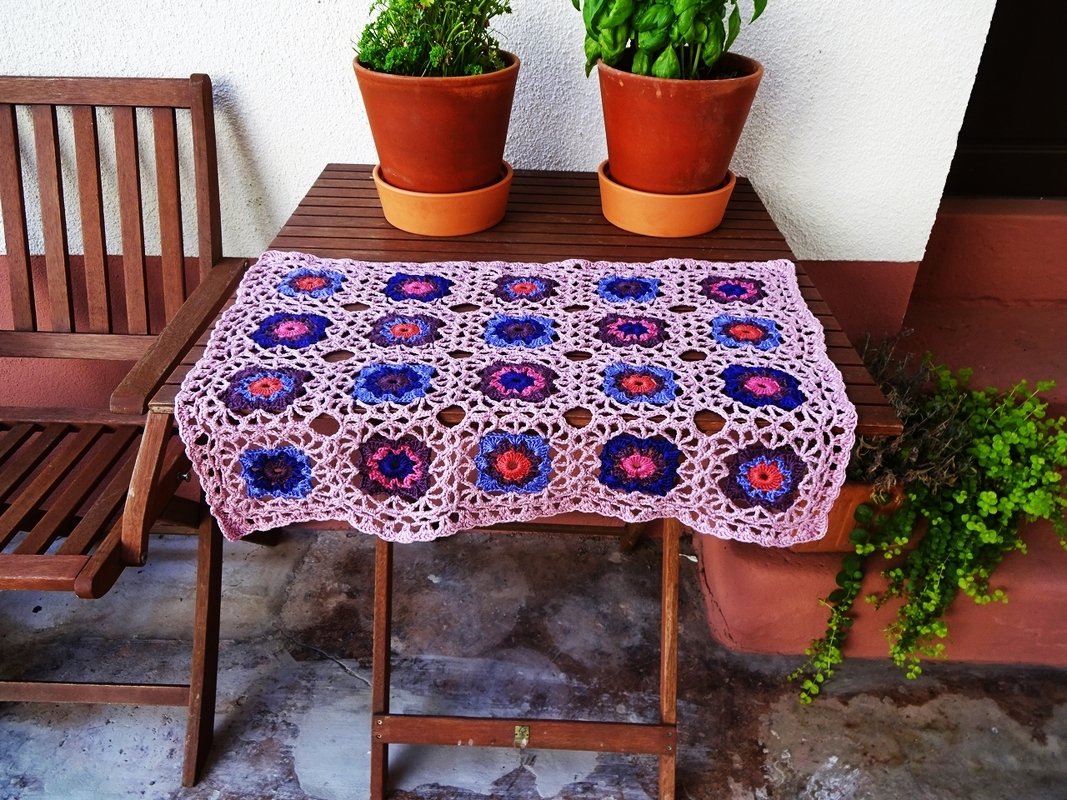 Gehäkelter Tischläufer aus verbundenen Blumenquadraten in Blau, Pink und Rot auf einem Holzklapptisch.