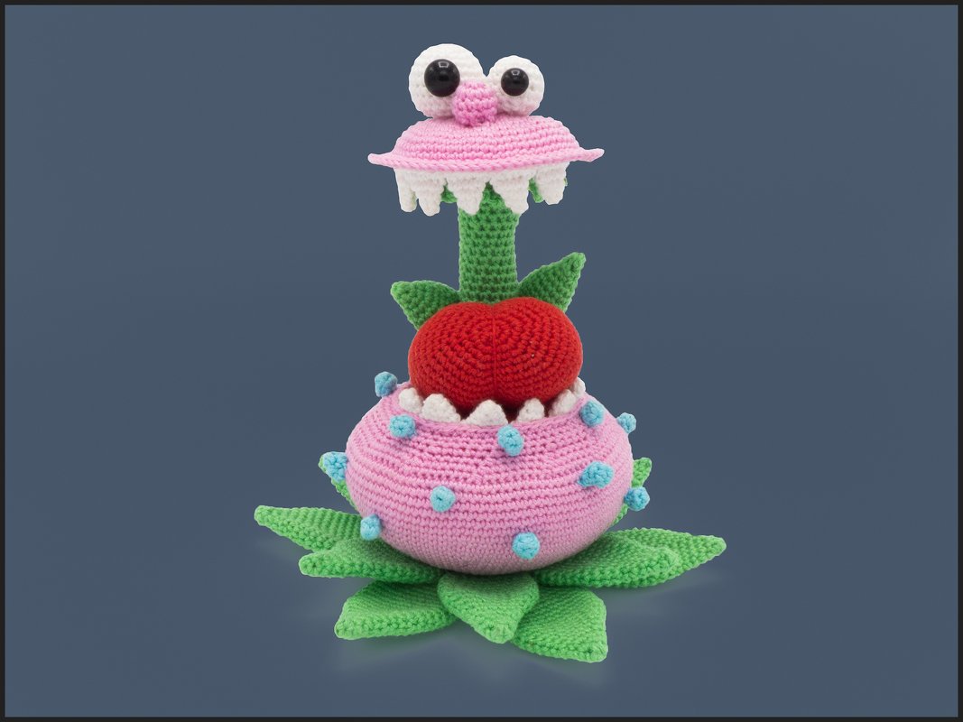 Gehäkelte rosa‑grüne Monsterpflanze mit großen Augen, weißen Zähnen und rotem Einsatz auf dunkelblauem Hintergrund.