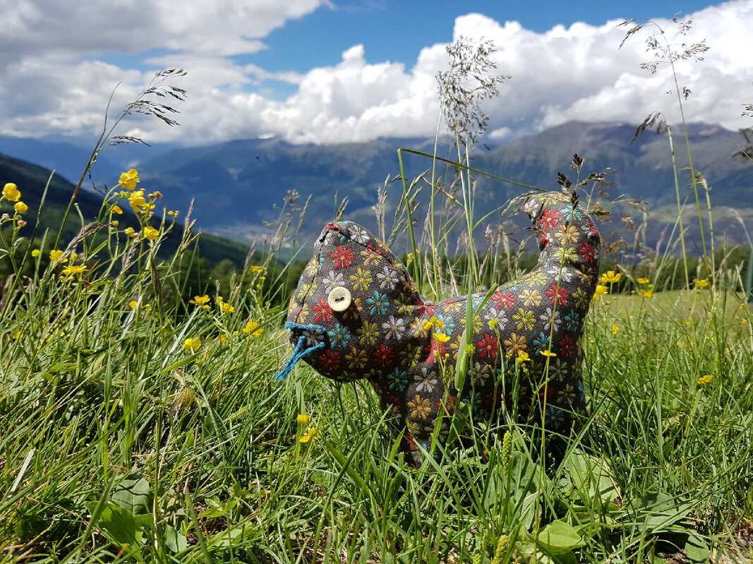 Genähtes Kuscheltier in Katzenform aus geblümtem Stoff mit Knopfauge und blauer Garnschnur in grüner Bergwiese vor Bergen