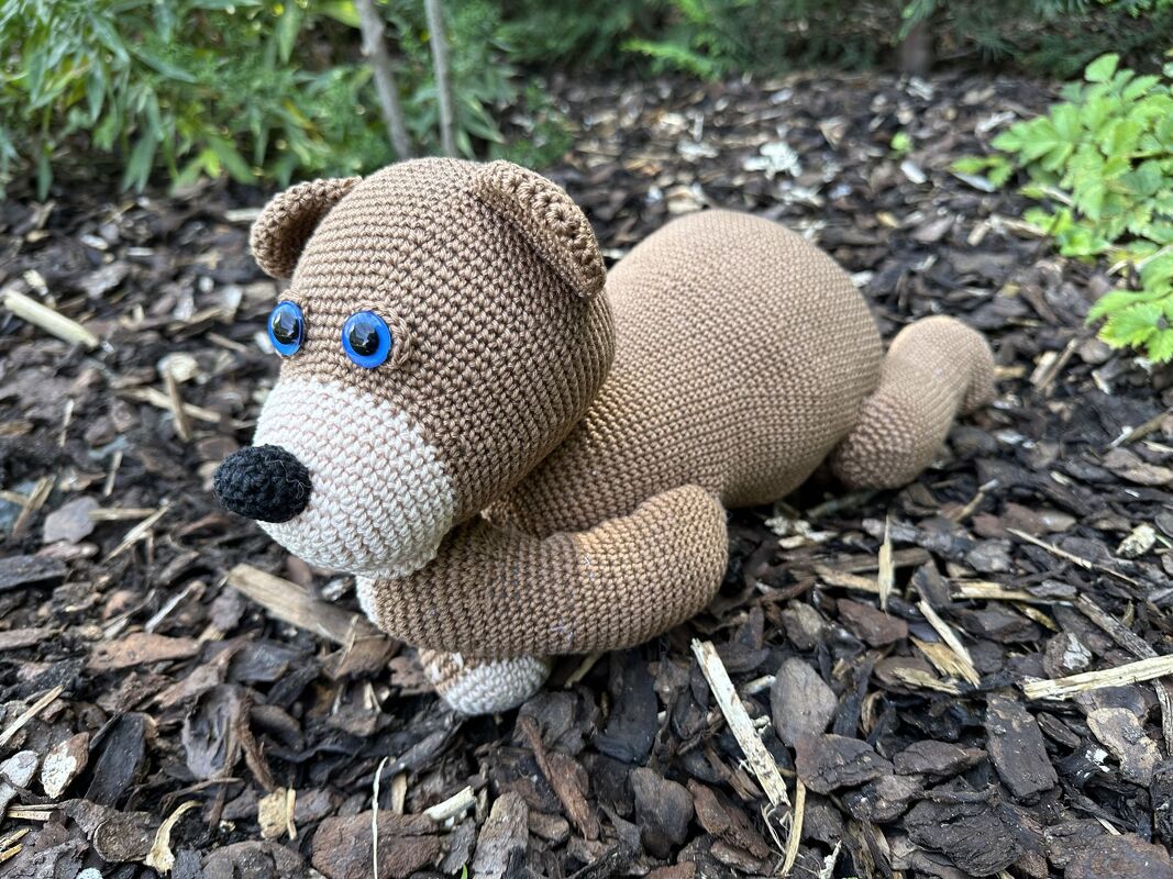 Brauner Amigurumi-Teddybär mit heller Schnauze und blauen Augen auf Rindenmulch