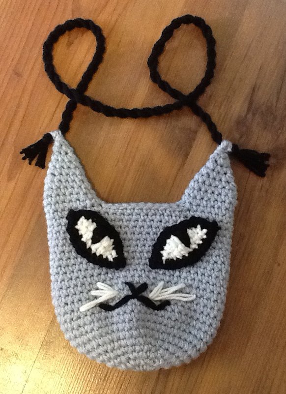 Hellgraue gehäkelte Umhängetasche in Katzenform mit schwarzem geflochtenem Trageband, applizierten Augen und weißen Schnurrhaaren.