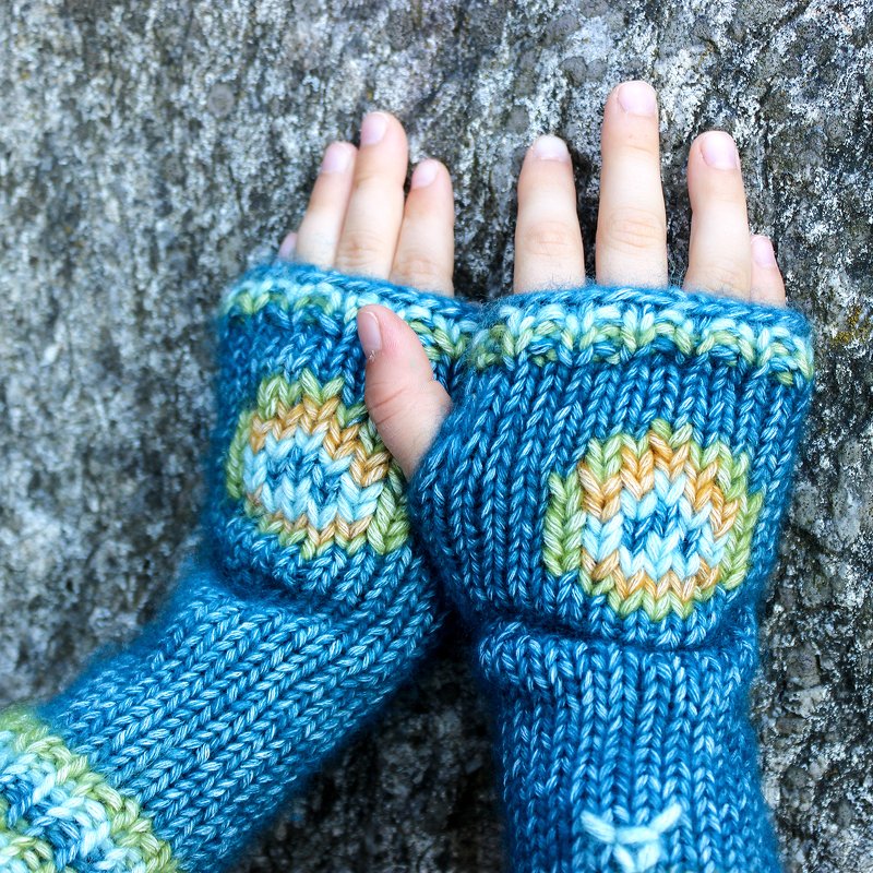 Gestrickte blaue fingerlose Kinder-Armstulpen mit rundem mehrfarbigem Motiv auf dem Handrücken, Hände vor Felswand