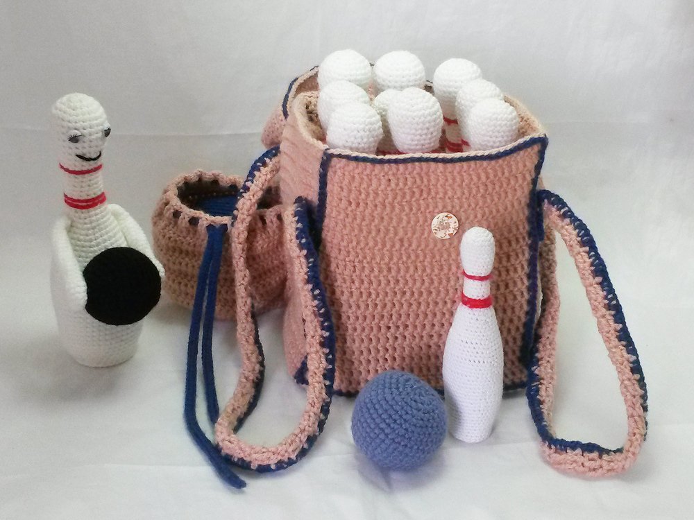 Gehäkeltes Kegelspiel: hellrosa Tasche mit dunkelblauer Umrandung, mehreren weißen Kegeln mit roten Ringen, einer blauen Häkelkugel und einem fröhlichen Kegel mit Gesicht.