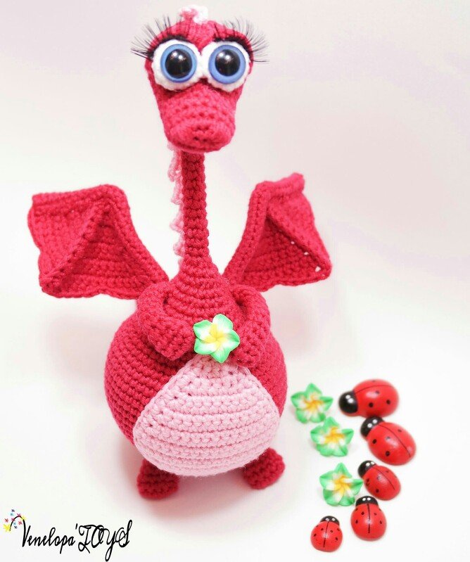 Red crocheted amigurumi dragon with large round eyes and a small flower on its chest