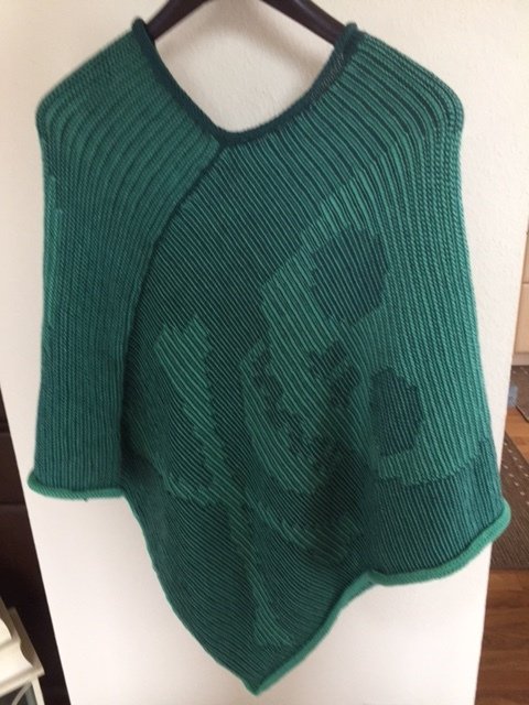 Grüner Strickponcho mit Rippenstruktur und dunklem Motiv, an einem Bügel hängend