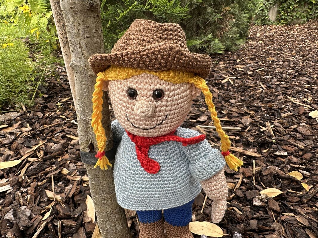 Gehäkelte Amigurumi-Puppe mit blonden Zöpfen, braunem Hut und rotem Halstuch, an einem Baumstamm gelehnt.