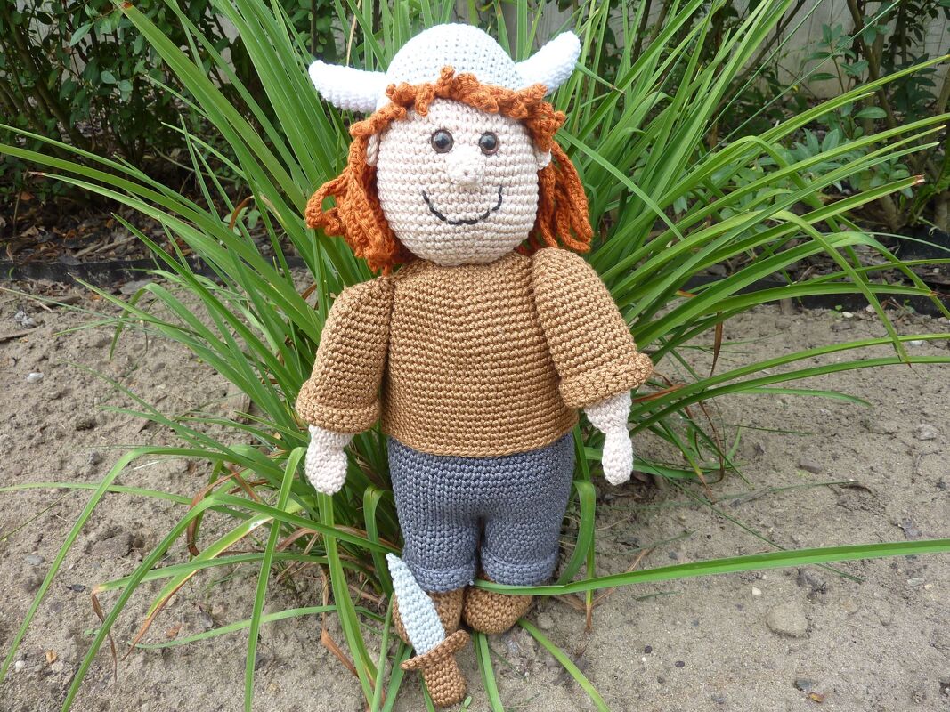Gehäkelte Amigurumi-Puppe mit Hörnerhelm, braunem Oberteil, grauen Hosen und kleinem Dolch vor grünen Pflanzen