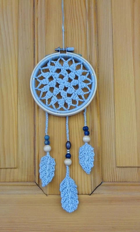 Hellblauer gehäkelter Traumfänger im Stickrahmen mit drei hängenden Häkelblättern und Holzperlen vor Holztür