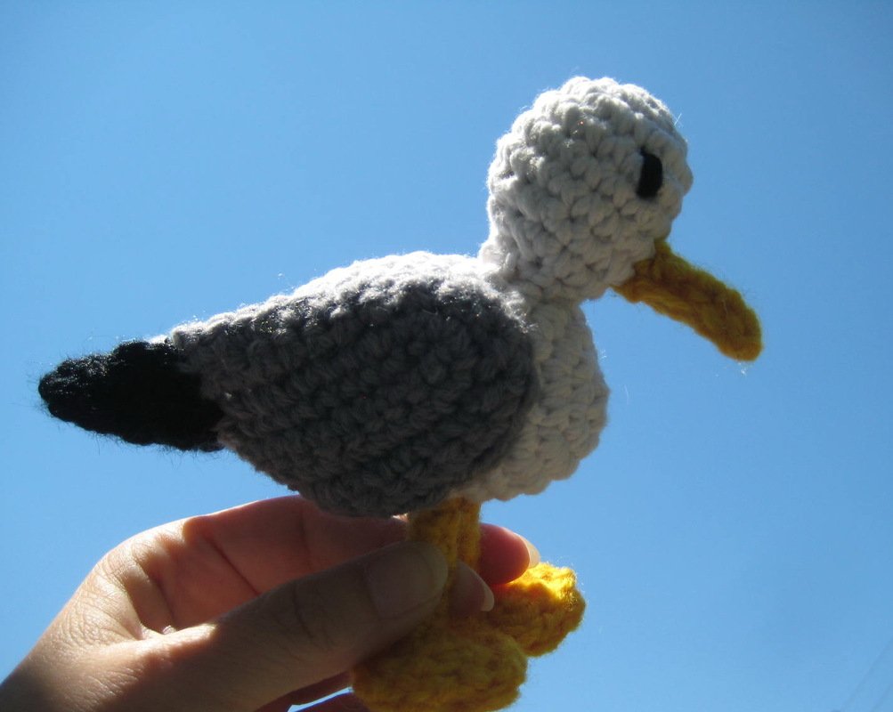 Grey and white crocheted amigurumi seagull with yellow beak held against blue sky