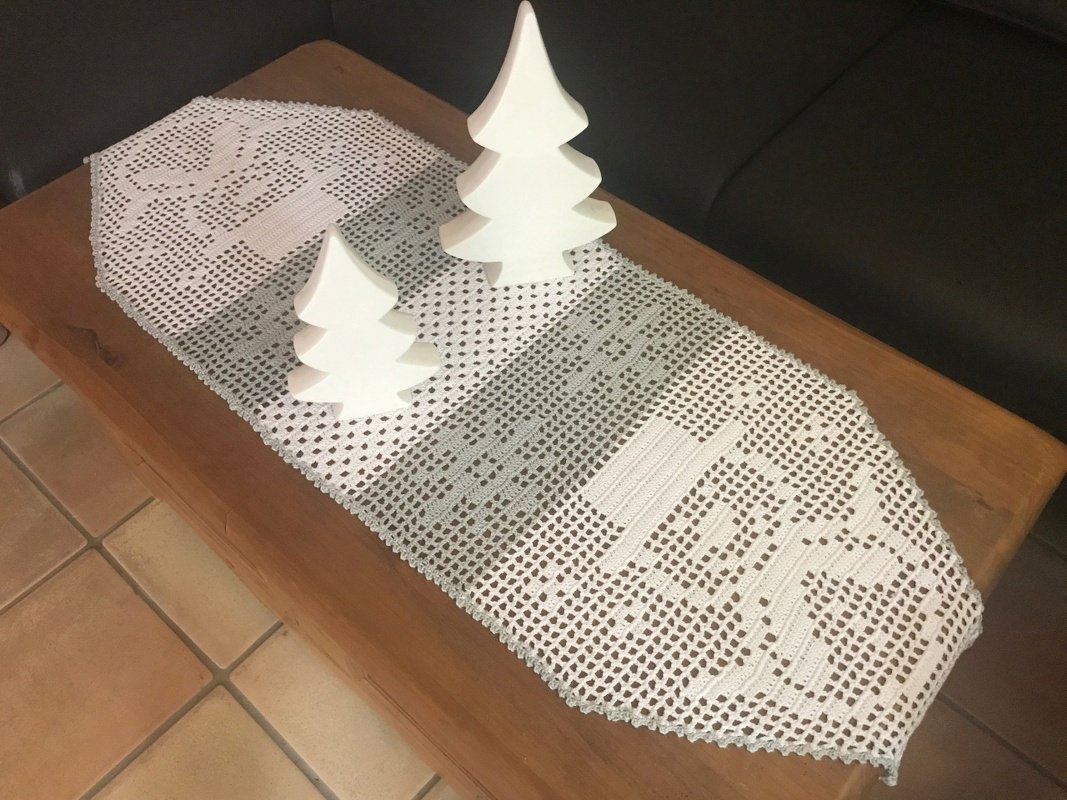 Weißer gehäkelter Tischläufer mit Rentiermotiv und zwei weißen Tannenbaumfiguren auf Holztisch